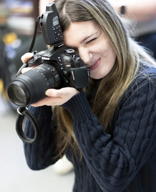 Student aiming a Nikon DSLR camera, squinting through the viewfinder in a well-lit studio space. Student aiming a Nikon DSLR camera, squinting through the viewfinder in a well-lit studio space.